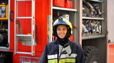 Portrait of happy female fireguard against the background of big red truck. Young firewoman in full equipment looking at camera with positive emotions near a fire engine. Concept of heroic profession. clipart