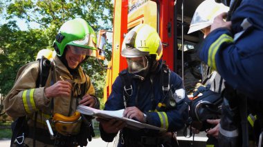 Koruyucu üniformalı erkek itfaiyeciler itfaiye aracının yanındaki kağıt talimatlarına bakarak kurtarma planını tartışıyor. Genç itfaiyeciler büyük kırmızı arabanın arka planındaki yangını söndürmeye hazır olun. Yavaş