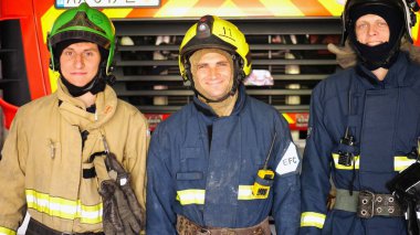 Korumalı üniformalı genç itfaiyecilerin portresi istasyondaki itfaiye aracının arka planında. Tam teçhizatlı erkek itfaiyeciler büyük kırmızı arabanın yanında pozitif duygularla kameraya bakıyorlar..