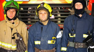 Büyük kırmızı bir arabanın yanında duran kasklı ve koruyucu üniformalı erkek itfaiyecilerin portresi. Tam teçhizatlı genç itfaiyeciler istasyondaki itfaiye arabasının arka planında poz veriyorlar..