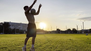 Genç adam gün batımında stadyumda futbol topuyla hokkabazlık yapıyor. Yeşil sahada profesyonel futbolcu topa vuruyor. Sporcu arka planda güneş ışığıyla çayırda numaralar yapıyor. Serbest stil futbol.