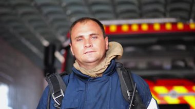 Departmandaki itfaiye arabasının yanında duran yorgun erkek itfaiyecinin portresi. Koruyucu üniformalı genç itfaiyeci büyük kırmızı kamyonun arka planındaki yangın üstüne kameraya bakıyor..