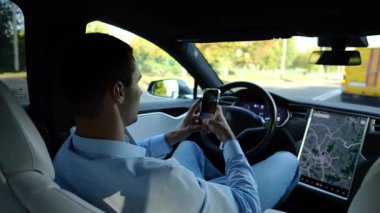 Başarılı bir iş adamı, otonom elektrikli araba kullanırken modern akıllı telefona göz atıyor. Şehir yolunda otomatik pilotla elektrikli araba sürerken telefon kullanan erkek girişimci..