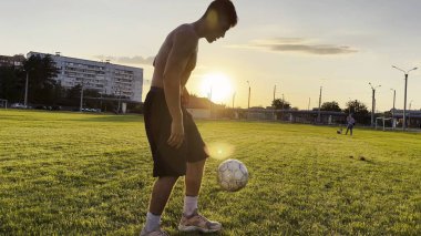 Genç adam gün batımında stadyumda futbol topuyla hokkabazlık yapıyor. Yeşil sahada profesyonel futbolcu topa vuruyor. Sporcu arka planda güneş ışığıyla çayırda numaralar yapıyor. Serbest stil futbol.