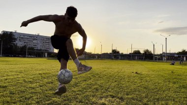 Genç adam gün batımında stadyumda futbol topuyla hokkabazlık yapıyor. Yeşil sahada profesyonel futbolcu topa vuruyor. Sporcu arka planda güneş ışığıyla çayırda numaralar yapıyor. Serbest stil futbol.