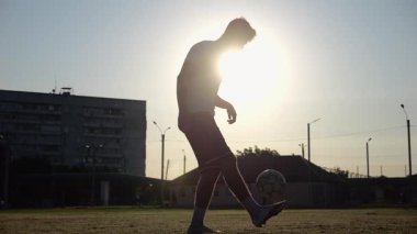 Günbatımında stadyumda futbol topu hokkabazlığı yapan profesyonel bir futbolcu. Genç adam yeşil sahada topa vuruyor. Sporcu arka planda güneş ışığıyla çayırda numaralar yapıyor. Serbest stil futbol kavramı.