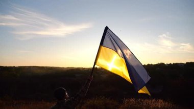 Erkek Ukrayna askeri, arkasında günbatımı olan bir Ukrayna bayrağı kaldırdı. Kamuflaj üniformalı genç adam, Rus saldırısına karşı kazanılan zaferin şerefine mavi-sarı bayrak kaldırdı..