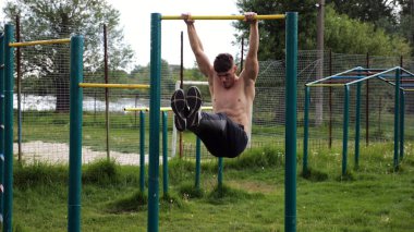 Atletik adam karın kaslarını eğitir spor sahasında yatay barda bacak kaldırır. Genç sporcu, barfiks çekme çubuğunda egzersiz yapıyor. Doğada spor yapan sporcu. Sağlıklı yaşam tarzı.