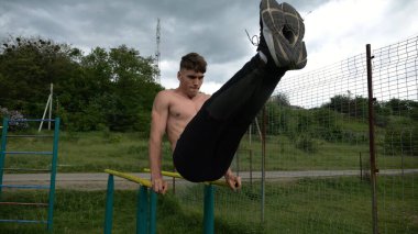 Güçlü bir adam karın kaslarını eğitir spor sahasında paralel barlarda bacak kaldırır. Genç üstsüz sporcu doğada egzersiz yapıyor. Doğada spor yapan sporcu. Sportif yaşam tarzı kavramı.