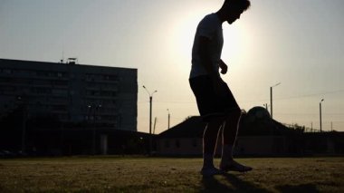 Günbatımında stadyumda futbol topu hokkabazlığı yapan profesyonel bir futbolcu. Genç adam yeşil sahada topa vuruyor. Sporcu arka planda güneş ışığıyla çayırda numaralar yapıyor. Serbest stil futbol kavramı.