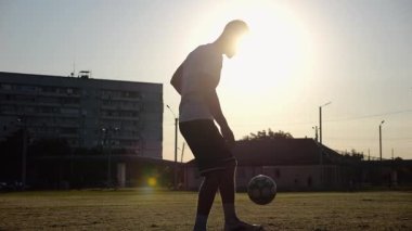 Günbatımında stadyumda futbol topu hokkabazlığı yapan profesyonel bir futbolcu. Genç adam yeşil sahada topa vuruyor. Sporcu arka planda güneş ışığıyla çayırda numaralar yapıyor. Serbest stil futbol kavramı.