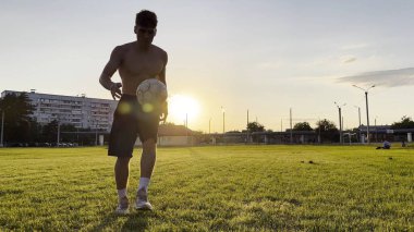 Genç adam gün batımında stadyumda futbol topuyla hokkabazlık yapıyor. Yeşil sahada profesyonel futbolcu topa vuruyor. Sporcu arka planda güneş ışığıyla çayırda numaralar yapıyor. Serbest stil futbol.