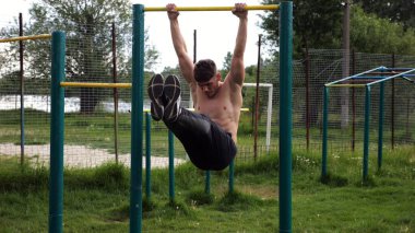 Atletik adam karın kaslarını eğitir spor sahasında yatay barda bacak kaldırır. Genç sporcu, barfiks çekme çubuğunda egzersiz yapıyor. Doğada spor yapan sporcu. Sağlıklı yaşam tarzı.