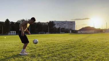 Genç adam gün batımında stadyumda futbol topuyla hokkabazlık yapıyor. Yeşil sahada profesyonel futbolcu topa vuruyor. Sporcu arka planda güneş ışığıyla çayırda numaralar yapıyor. Serbest stil futbol.
