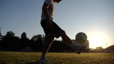 Günbatımında stadyumda futbol topu hokkabazlığı yapan profesyonel bir futbolcu. Genç adam yeşil sahada topa vuruyor. Sporcu arka planda güneş ışığıyla çayırda numaralar yapıyor. Serbest stil futbol kavramı.