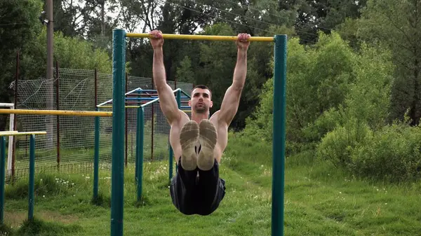 Atletik adam karın kaslarını eğitir spor sahasında yatay barda bacak kaldırır. Genç sporcu, barfiks çekme çubuğunda egzersiz yapıyor. Doğada spor yapan sporcu. Sağlıklı yaşam tarzı.