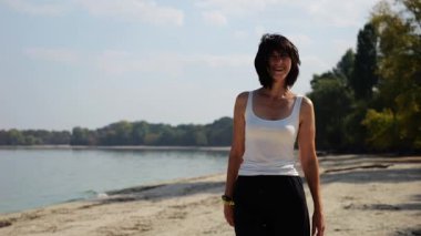 Güneşli bir günde sahil kenarında kumsalda yürüyen güzel bir bayan. Mutlu yetişkin kadın kıyı şeridine doğru dönüyor. Kaygısız kadın deniz kenarında dinleniyor. Yaz tatili veya tatil kavramı.