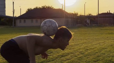 Genç adam gün batımında stadyumda futbol topuyla hokkabazlık yapıyor. Yeşil sahada profesyonel futbolcu topa vuruyor. Sporcu arka planda güneş ışığıyla çayırda numaralar yapıyor. Serbest stil futbol.