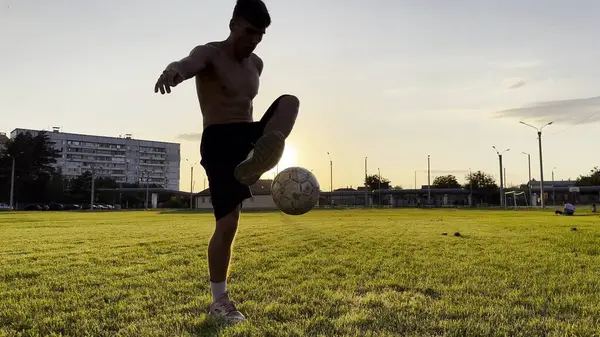 Genç adam gün batımında stadyumda futbol topuyla hokkabazlık yapıyor. Yeşil sahada profesyonel futbolcu topa vuruyor. Sporcu arka planda güneş ışığıyla çayırda numaralar yapıyor. Serbest stil futbol.