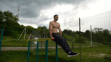 Güçlü bir adam karın kaslarını eğitir spor sahasında paralel barlarda bacak kaldırır. Genç üstsüz sporcu doğada egzersiz yapıyor. Doğada spor yapan sporcu. Sportif yaşam tarzı kavramı.