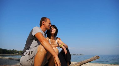 Kumsalda tahta kütüğün üzerinde otururken mutlu çift kucaklaşması. Güneşli bir günde deniz manzarasına hayran olan romantik bir çift. Sevgililer tatil köyünde birlikte vakit geçirirler. Yaz tatili ya da tatil kavramı. Yavaş çekim