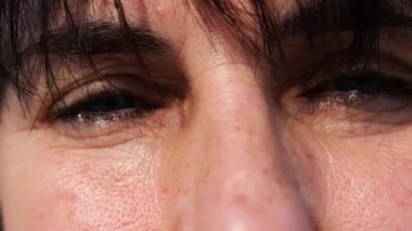 Gözünü kırpıp dışarıdaki kameraya bakan bir kadının yorgun mavi gözleri. Güneşin parlak ışıklarıyla aydınlatan yetişkin bir kadının yüzü. Hanımefendinin üzgün yüz ifadesi. Yavaşla.