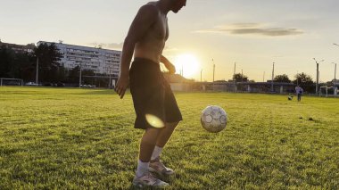 Genç adam gün batımında stadyumda futbol topuyla hokkabazlık yapıyor. Yeşil sahada profesyonel futbolcu topa vuruyor. Sporcu arka planda güneş ışığıyla çayırda numaralar yapıyor. Serbest stil futbol.