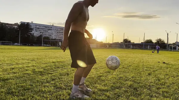Genç adam gün batımında stadyumda futbol topuyla hokkabazlık yapıyor. Yeşil sahada profesyonel futbolcu topa vuruyor. Sporcu arka planda güneş ışığıyla çayırda numaralar yapıyor. Serbest stil futbol.