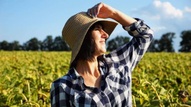 Mutlu gülümseyen dişi çiftçi ayçiçeği tarlasına bakar ve hasadın tadını çıkarır. Hasır şapkalı, sarı tarlada yetişmiş güzel tarımcı portresi. Tarım sektörü kavramı.