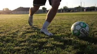 Profesyonel futbolcunun erkek ayağı, yeşil sahada koşarken topla numaralar gösterir. Sporcu bacakları gün batımında çayırda koşarken topa vurur. Stadyumda antrenman yapan genç adam. Yavaş çekim