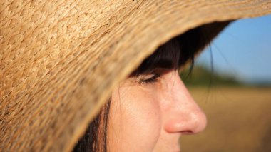 Hasır şapkalı kadın çiftçi buğday tarlasına bakıyor ve gün batımında çiftliği inceliyor. Güneşin parlak ışıklarıyla aydınlatan yetişkin ve güzel bir kadının yüzünü kapatın. Tarım sektörü kavramı.