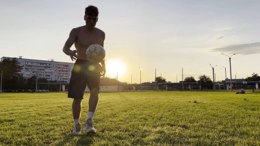 Genç adam gün batımında stadyumda futbol topuyla hokkabazlık yapıyor. Yeşil sahada profesyonel futbolcu topa vuruyor. Sporcu arka planda güneş ışığıyla çayırda numaralar yapıyor. Serbest stil futbol.