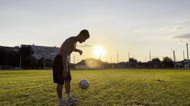 Genç adam gün batımında stadyumda futbol topuyla hokkabazlık yapıyor. Yeşil sahada profesyonel futbolcu topa vuruyor. Sporcu arka planda güneş ışığıyla çayırda numaralar yapıyor. Serbest stil futbol.