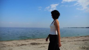 Güneşli bir günde sahilde kumsala giden mutlu yetişkin kadın. Arka planda sakin dalgalarla deniz kıyısında yürüyen güzel bir bayan. Yaz tatili ya da tatil kavramı. Yavaş çekim