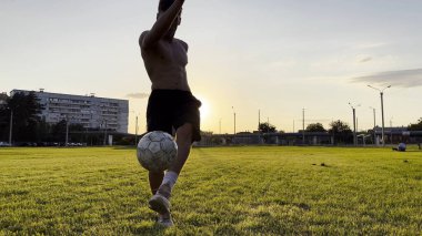 Genç adam gün batımında stadyumda futbol topuyla hokkabazlık yapıyor. Yeşil sahada profesyonel futbolcu topa vuruyor. Sporcu arka planda güneş ışığıyla çayırda numaralar yapıyor. Serbest stil futbol.