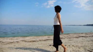 Güneşli bir günde sahilde kumsala giden mutlu yetişkin kadın. Arka planda sakin dalgalarla deniz kıyısında yürüyen güzel bir bayan. Yaz tatili ya da tatil kavramı. Yavaş çekim