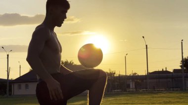 Genç adam gün batımında stadyumda futbol topuyla hokkabazlık yapıyor. Yeşil sahada profesyonel futbolcu topa vuruyor. Sporcu arka planda güneş ışığıyla çayırda numaralar yapıyor. Serbest stil futbol.