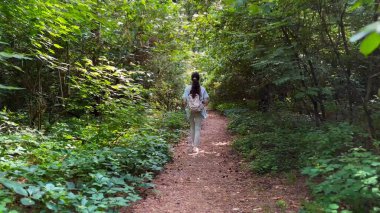 Genç bir kadın yaz ormanında patika boyunca gidiyor. Sırt çantalı bir kadın vahşi doğada patikaların arasında yürüyor. Tanınmayan bir kız dışarıda gezmekten zevk alıyor. Manzaraya hayran kalıyor. Kapat.