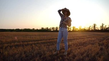 Hasır şapkalı yetişkin tarım uzmanı gün batımında çayırda arpa arıyor. Alacakaranlıkta olgun buğdayı inceleyen dişi çiftçi. Güzel manzara manzarası. Tarım ve tarım sektörü kavramı.