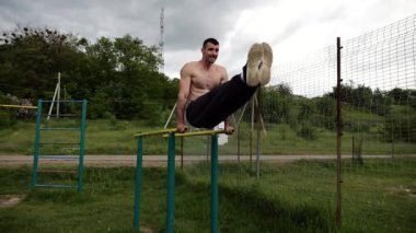 Güçlü bir adam karın kaslarını eğitir spor sahasında paralel barlarda bacak kaldırır. Genç üstsüz sporcu doğada egzersiz yapıyor. Doğada spor yapan sporcu. Sportif yaşam tarzı kavramı.