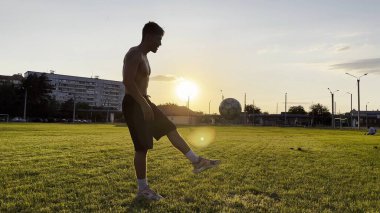 Genç adam gün batımında stadyumda futbol topuyla hokkabazlık yapıyor. Yeşil sahada profesyonel futbolcu topa vuruyor. Sporcu arka planda güneş ışığıyla çayırda numaralar yapıyor. Serbest stil futbol.