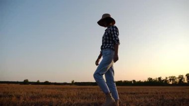 Alacakaranlıkta arpa tarlasında yürüyen hasır şapkalı yetişkin tarım uzmanı. Dişi çiftçi gün batımında buğday çayırından geçiyor. Güzel manzara manzarası. Tarım sektörü kavramı. Yavaş çekim