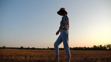 Alacakaranlıkta arpa tarlasında yürüyen hasır şapkalı yetişkin tarım uzmanı. Dişi çiftçi gün batımında buğday çayırından geçiyor. Güzel manzara manzarası. Tarım sektörü kavramı. Yavaş çekim