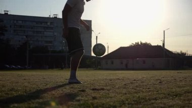 Günbatımında stadyumda futbol topu hokkabazlığı yapan profesyonel bir futbolcu. Genç adam yeşil sahada topa vuruyor. Sporcu arka planda güneş ışığıyla çayırda numaralar yapıyor. Serbest stil futbol kavramı.