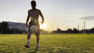 Genç adam gün batımında stadyumda futbol topuyla hokkabazlık yapıyor. Yeşil sahada profesyonel futbolcu topa vuruyor. Sporcu arka planda güneş ışığıyla çayırda numaralar yapıyor. Serbest stil futbol.