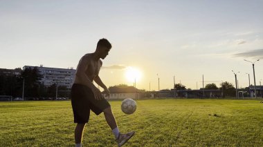 Genç adam gün batımında stadyumda futbol topuyla hokkabazlık yapıyor. Yeşil sahada profesyonel futbolcu topa vuruyor. Sporcu arka planda güneş ışığıyla çayırda numaralar yapıyor. Serbest stil futbol.