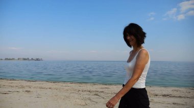 Güneşli bir günde sahilde kumsala giden mutlu yetişkin kadın. Arka planda sakin dalgalarla deniz kıyısında yürüyen güzel bir bayan. Yaz tatili ya da tatil kavramı. Yavaş çekim