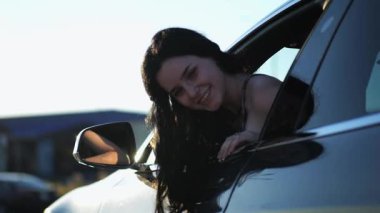 Genç, güzel bir kız, modern elektrikli arabaya doğru eğilmiş ve kameraya bakıyor. Alternatif elektrikli araçta gülümseyen çekici bir kadının portresi. Yavaş çekim