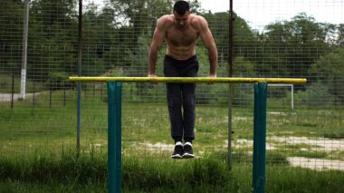 Cesur üstsüz sporcu doğadaki paralel parmaklıklar üzerinde egzersiz yapıyor. Spor alanında çalışan güçlü ve kaslı bir adam. Genç atletik adam dışarıda egzersiz yapıyor. Aktif sağlıklı yaşam tarzı kavramı.