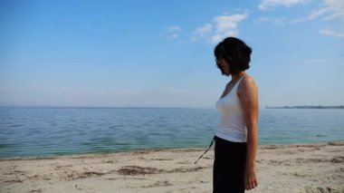 Güneşli bir günde sahilde kumsala giden mutlu yetişkin kadın. Arka planda sakin dalgalarla deniz kıyısında yürüyen güzel bir bayan. Yaz tatili ya da tatil kavramı. Yavaş çekim